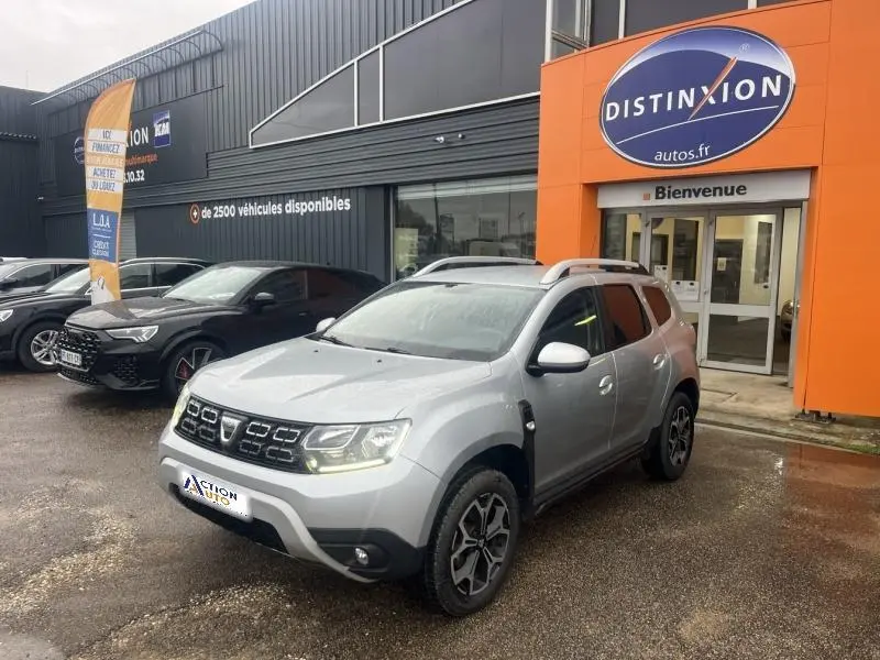 Dacia Duster gris Comète métallisé vu en 3/4 avant droit, avec barres de toit et jantes alu devant un concessionnaire.