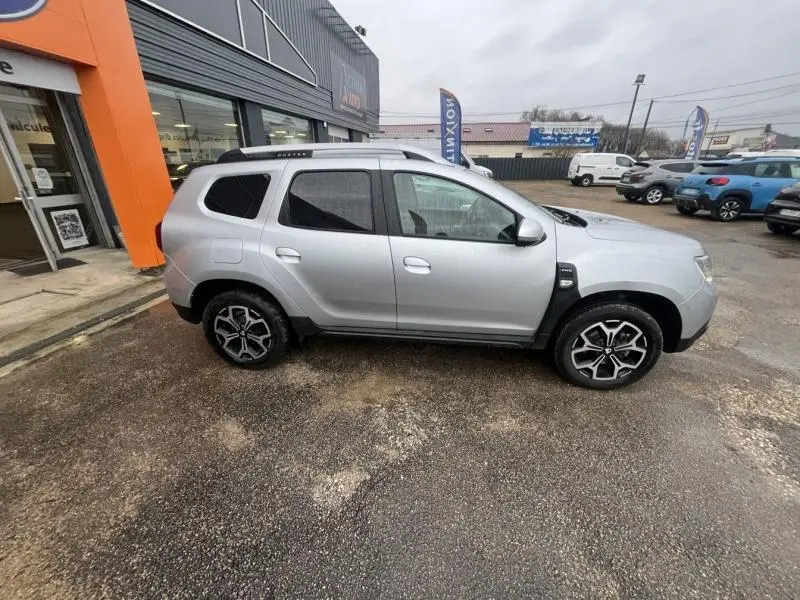 Profil côté droit du Dacia Duster gris Comète métallisé 2021 avec jantes alu et barres de toit visibles.
