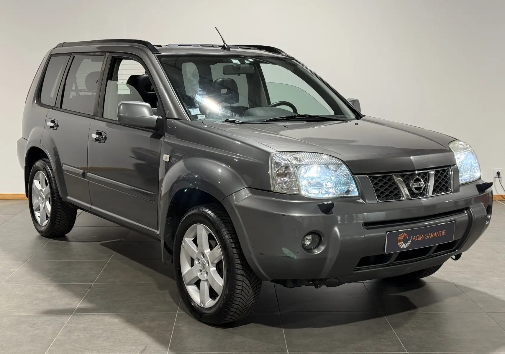 Nissan X-Trail Columbia gris vu en 3/4 avant droit, phares allumés et jantes alliage distinctives visibles.