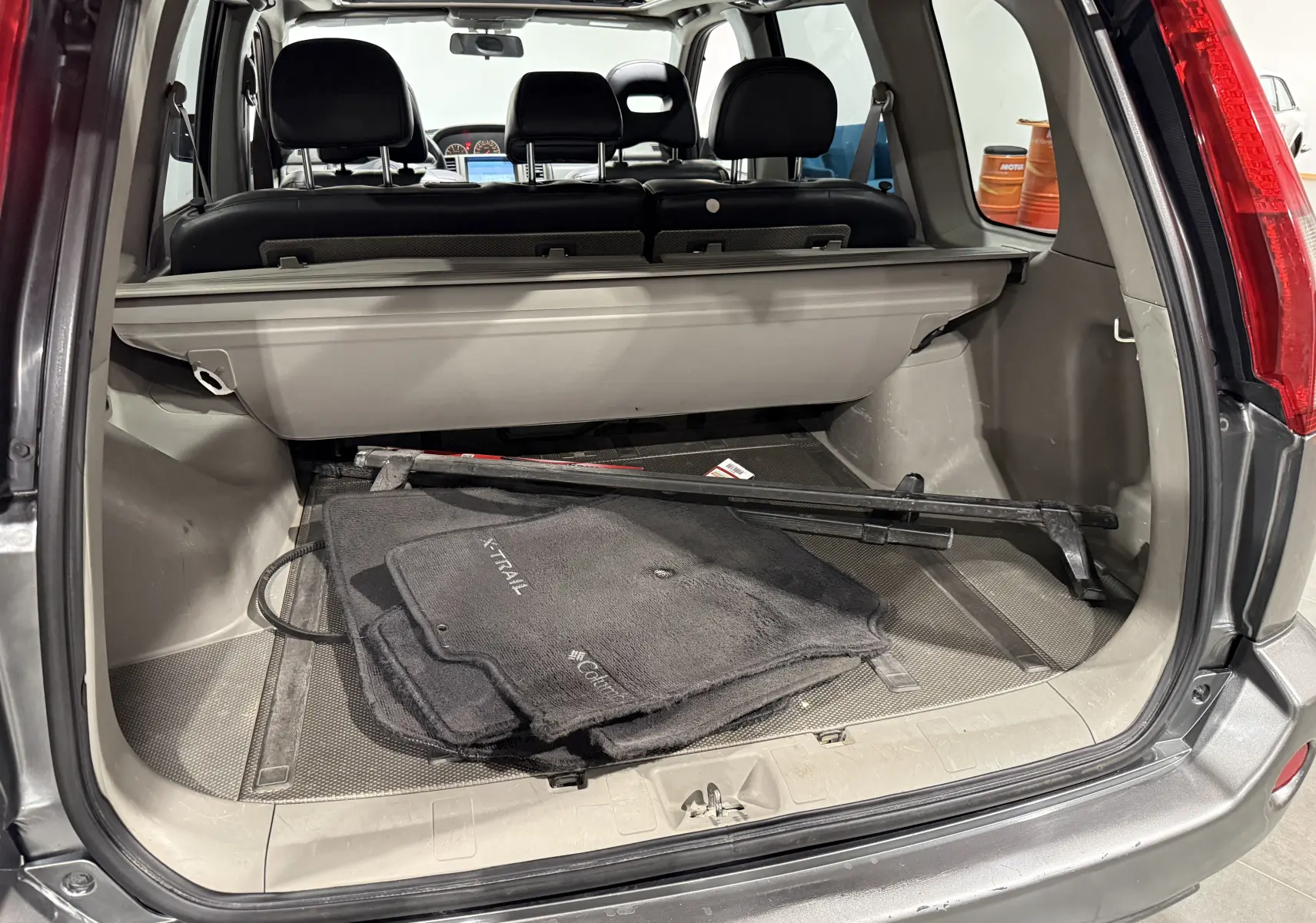 Vue arrière du coffre ouvert d’un Nissan X-Trail Columbia gris 2006 avec tapis de sol et cache-bagages visibles.