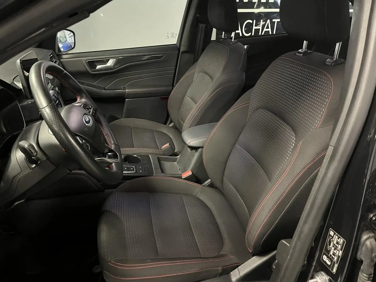 Intérieur avant du Ford Kuga noir 2022, sièges sport tissu noir avec surpiqûres rouges et volant ST-Line à méplat.