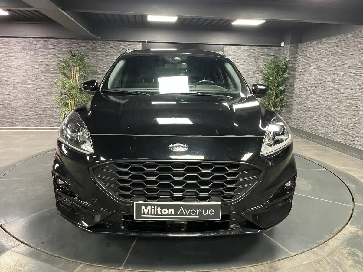 Vue frontale d'un Ford Kuga noir 2022 avec calandre hexagonale et badge Ford visible en intérieur showroom.