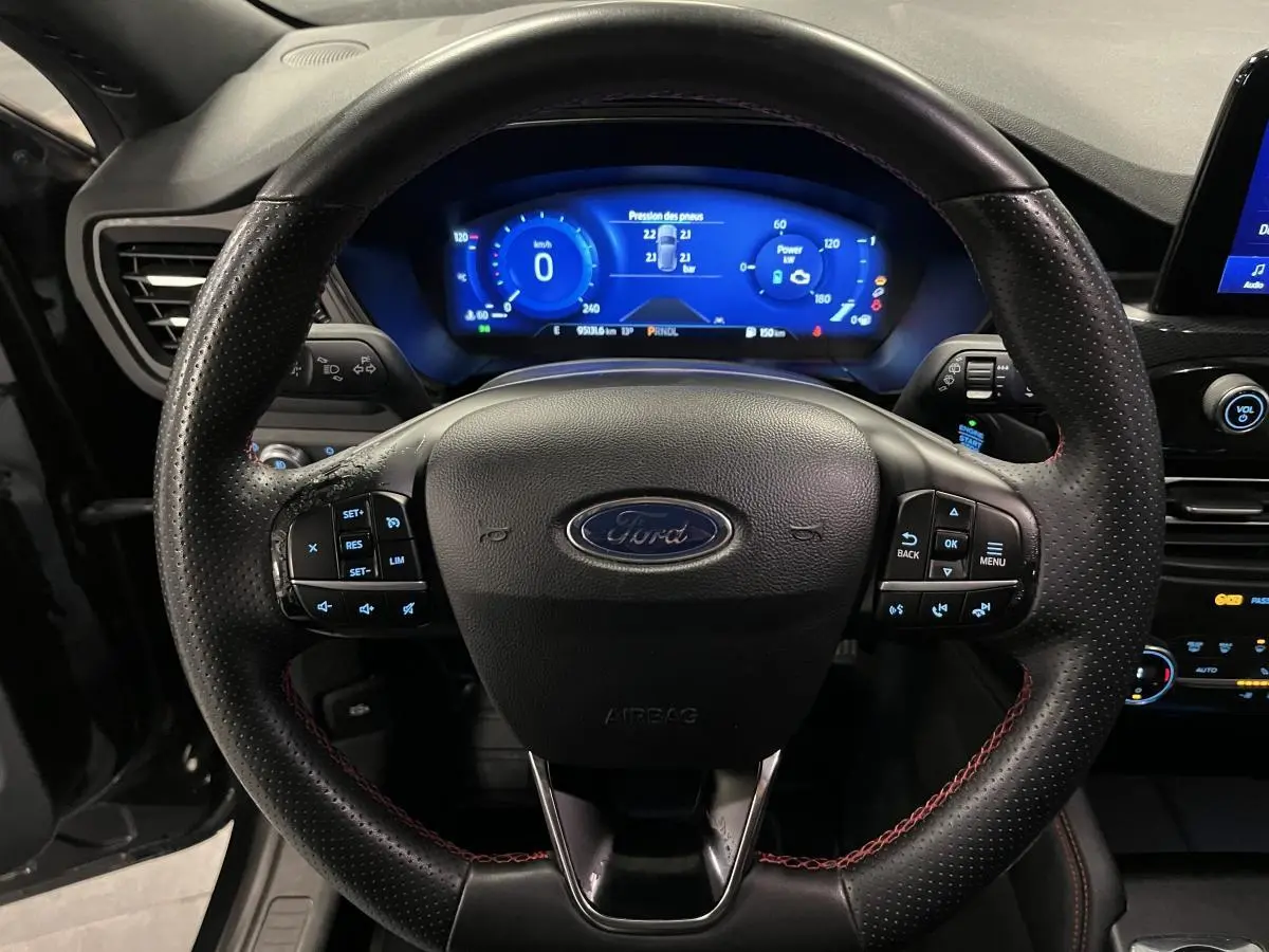 Vue rapprochée du volant noir perforé avec surpiqûres rouges et tableau de bord numérique bleu du Ford Kuga 2022 ST-Line.