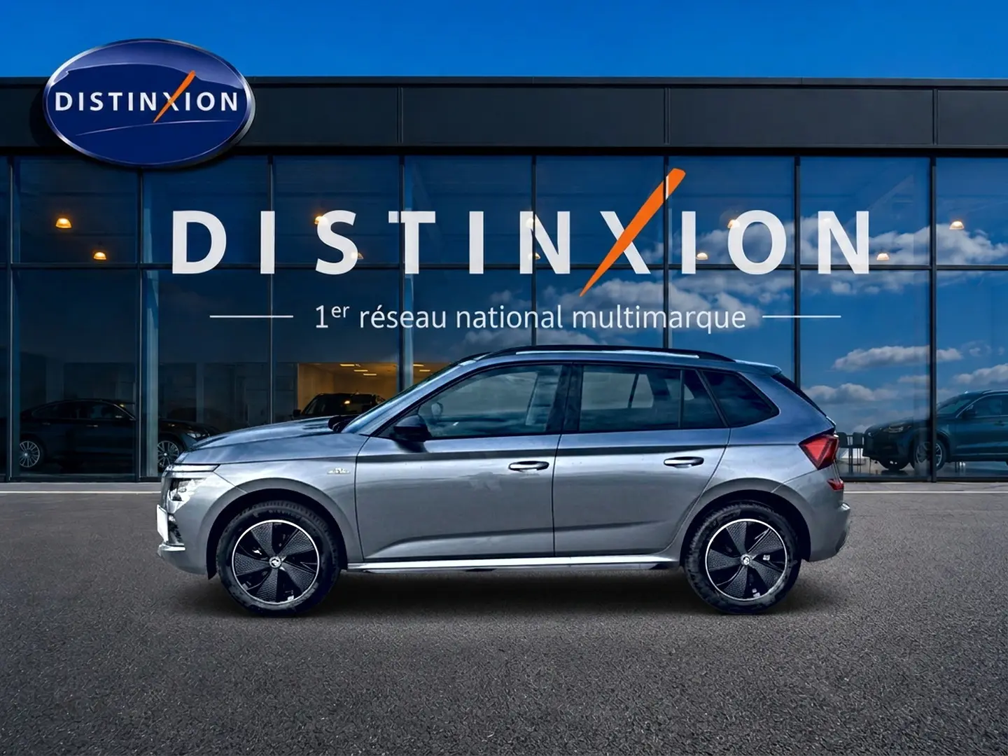 Vue latérale droite du Skoda Kamiq gris graphite avec jantes alu noires et toit panoramique en verre, devant une vitrine Distinxion.