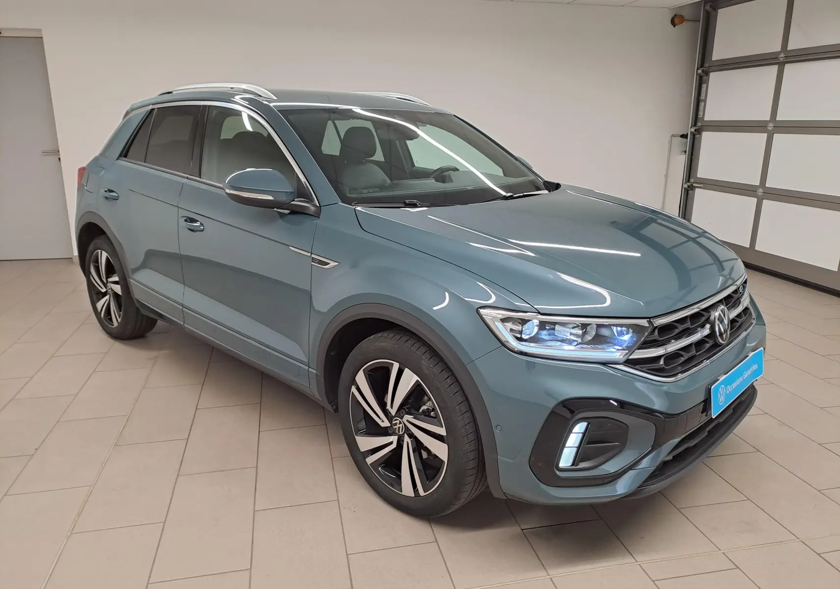 Volkswagen T-Roc 2025 bleu pétrole en 3/4 avant droit, avec phares allumés et jantes R-Line distinctives.