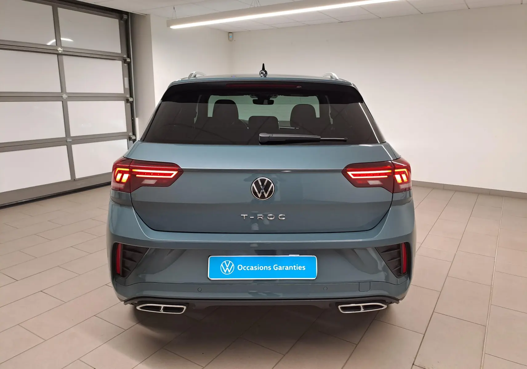 Vue arrière d'un Volkswagen T-Roc bleu pétrole 2025 avec feux LED allumés et double sortie d'échappement chromée.
