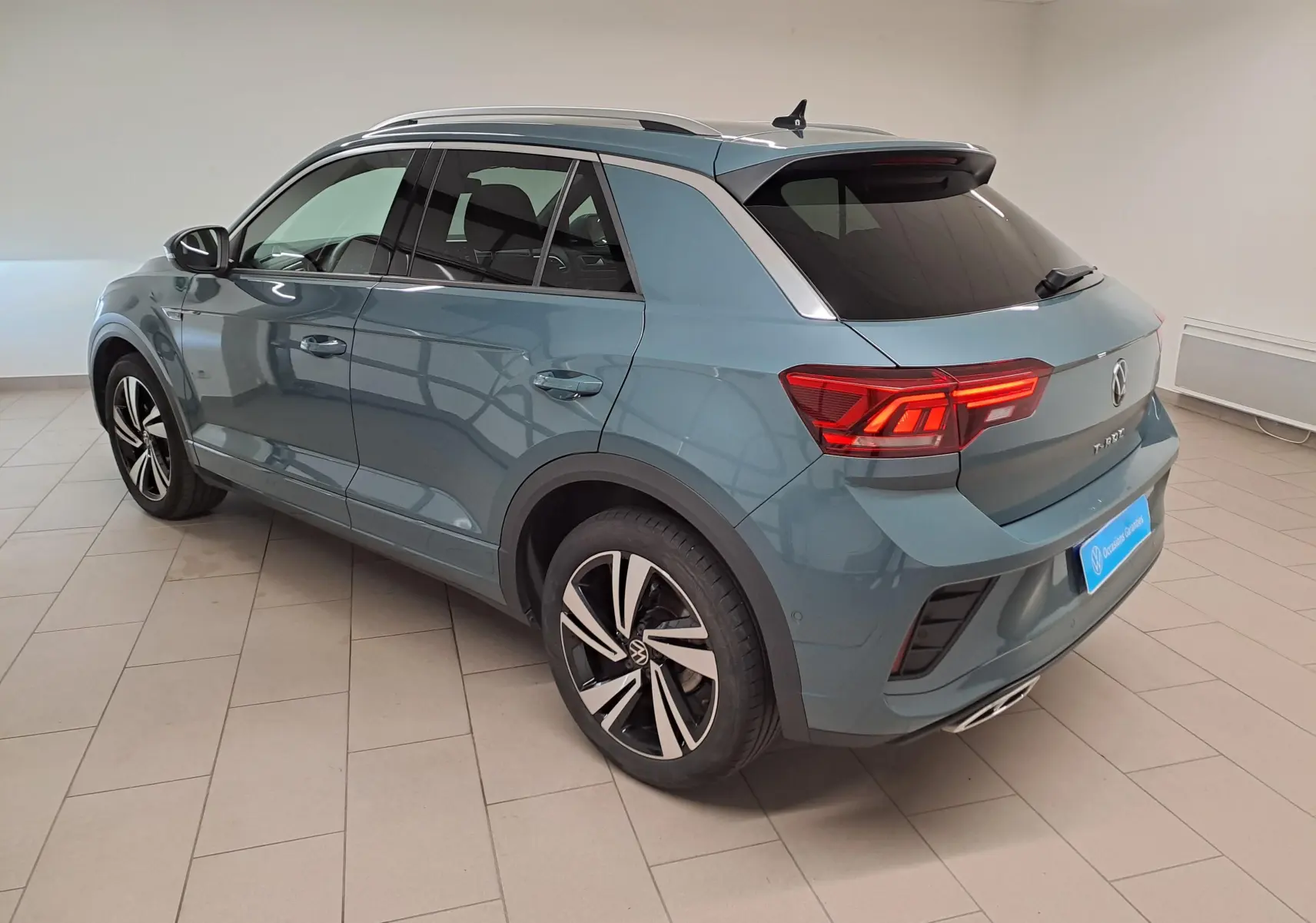 Volkswagen T-Roc 2025 bleu pétrole en vue 3/4 arrière droit, avec jantes alliage et feux arrière allumés.