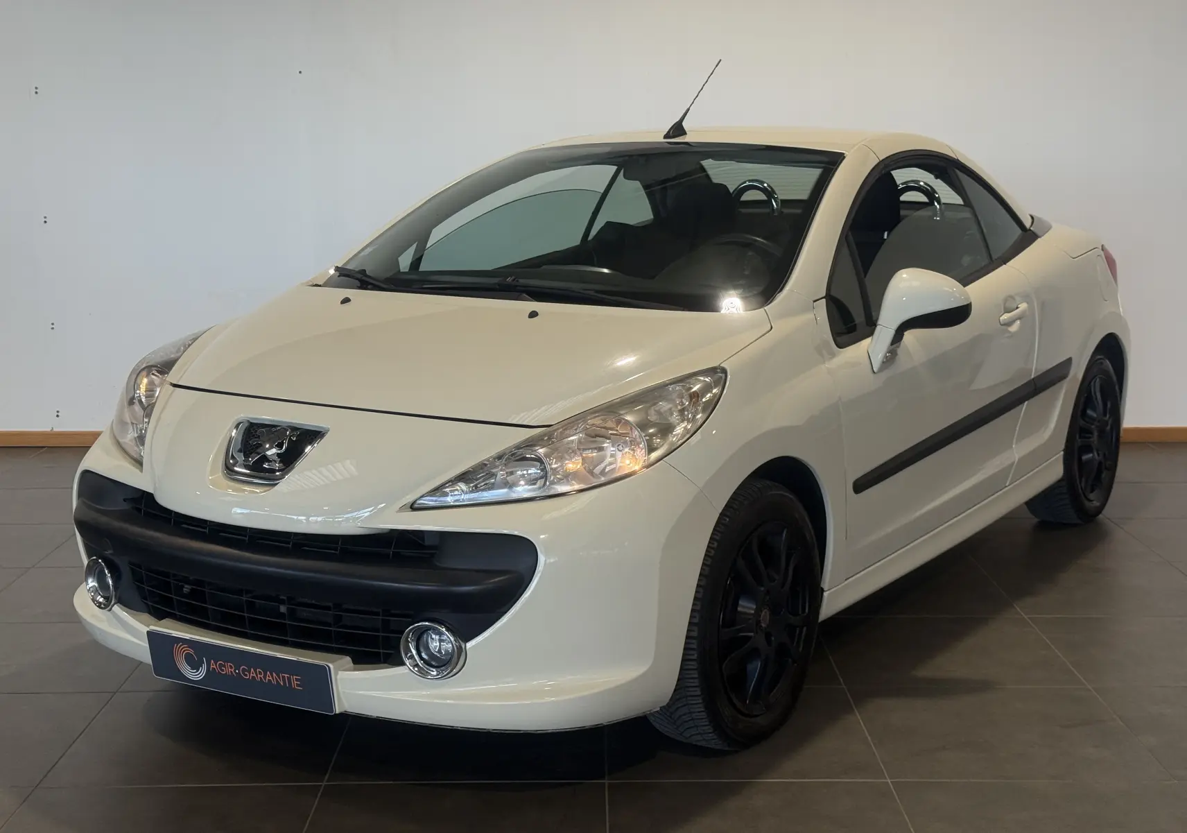 Peugeot 207 CC beige vue 3/4 avant droit, cabriolet fermé, jantes noires et phares allumés en intérieur.