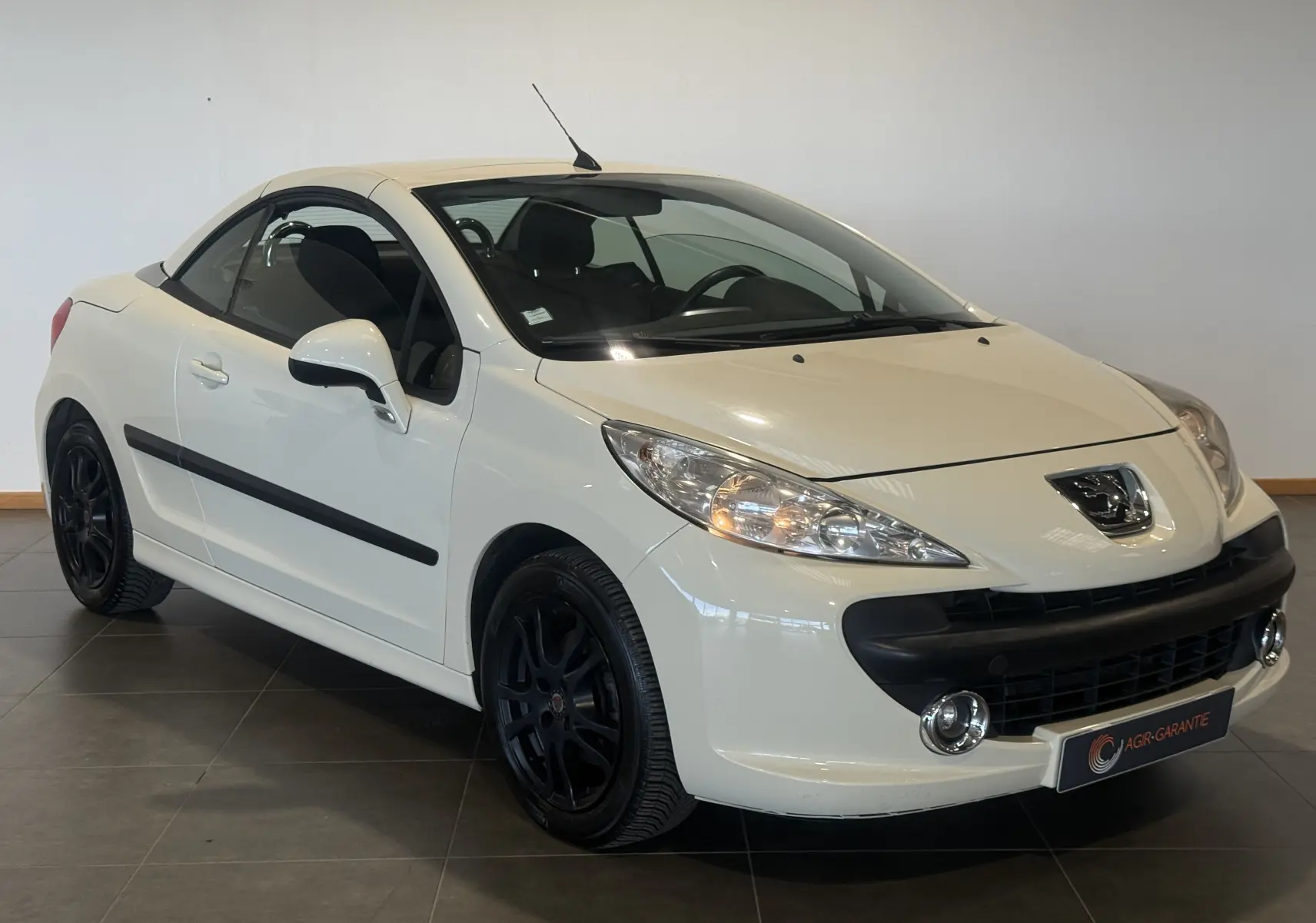 Peugeot 207 CC beige vue 3/4 avant droit, cabriolet fermé, jantes noires et phares allumés.