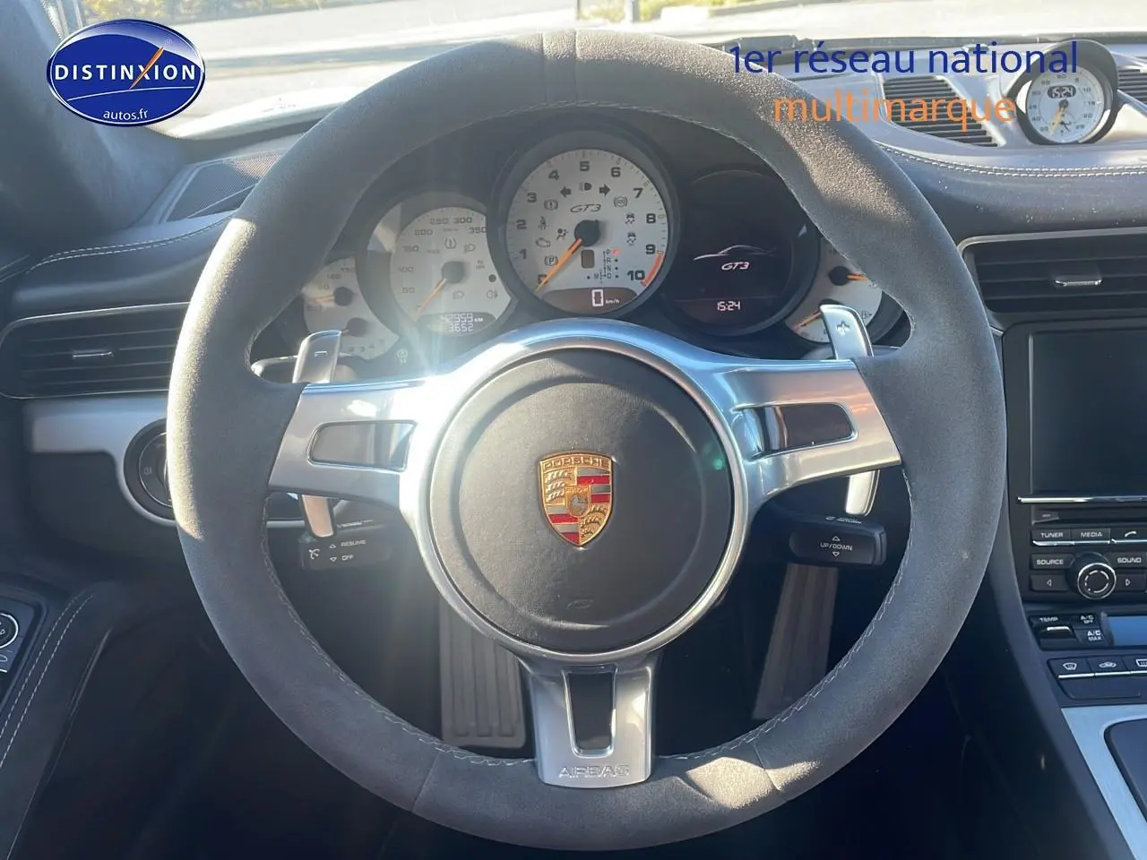 Vue intérieure centrée sur le volant en alcantara gris d’une Porsche 911 GT3 blanc, avec compte-tours et tableau de bord visibles.