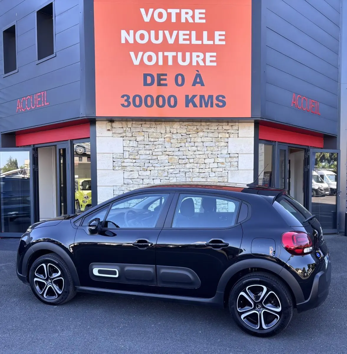 Profil latéral droit d'une Citroën C3 noire 2024 avec protections latérales caractéristiques, stationnée devant un bâtiment commercial.