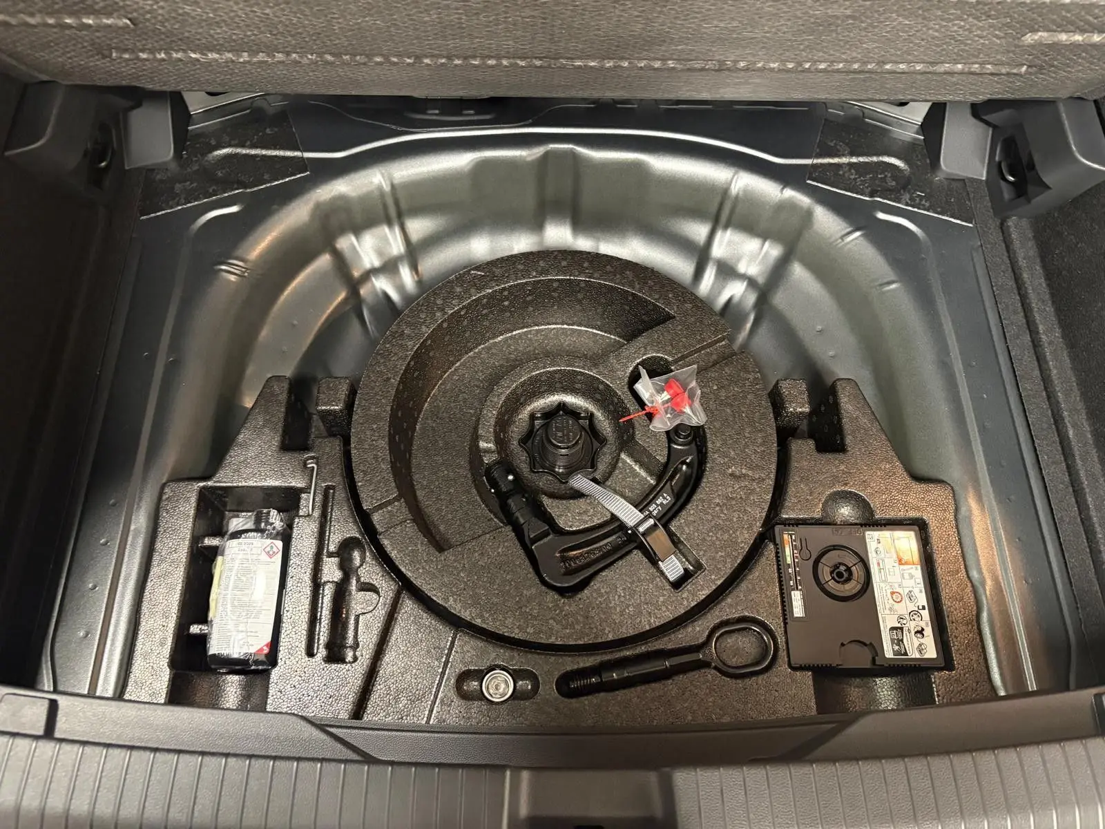 Vue du coffre ouvert du Volkswagen T-Roc 2025 gris Indium, montrant le rangement sous le plancher avec dispositif d'attelage amovible.