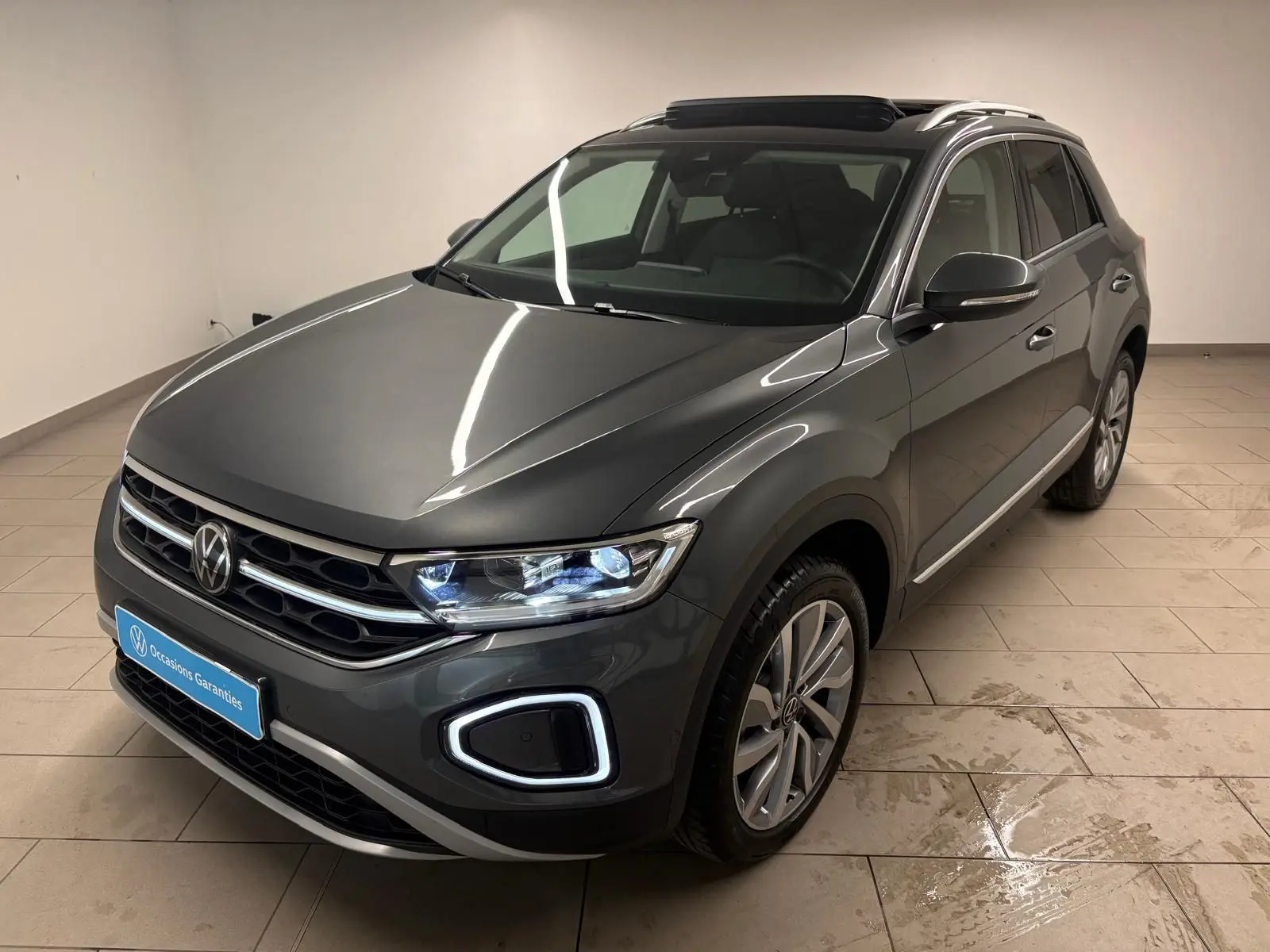 Volkswagen T-Roc 2025 gris Indium en 3/4 avant droit avec feux allumés et attelage amovible visible.