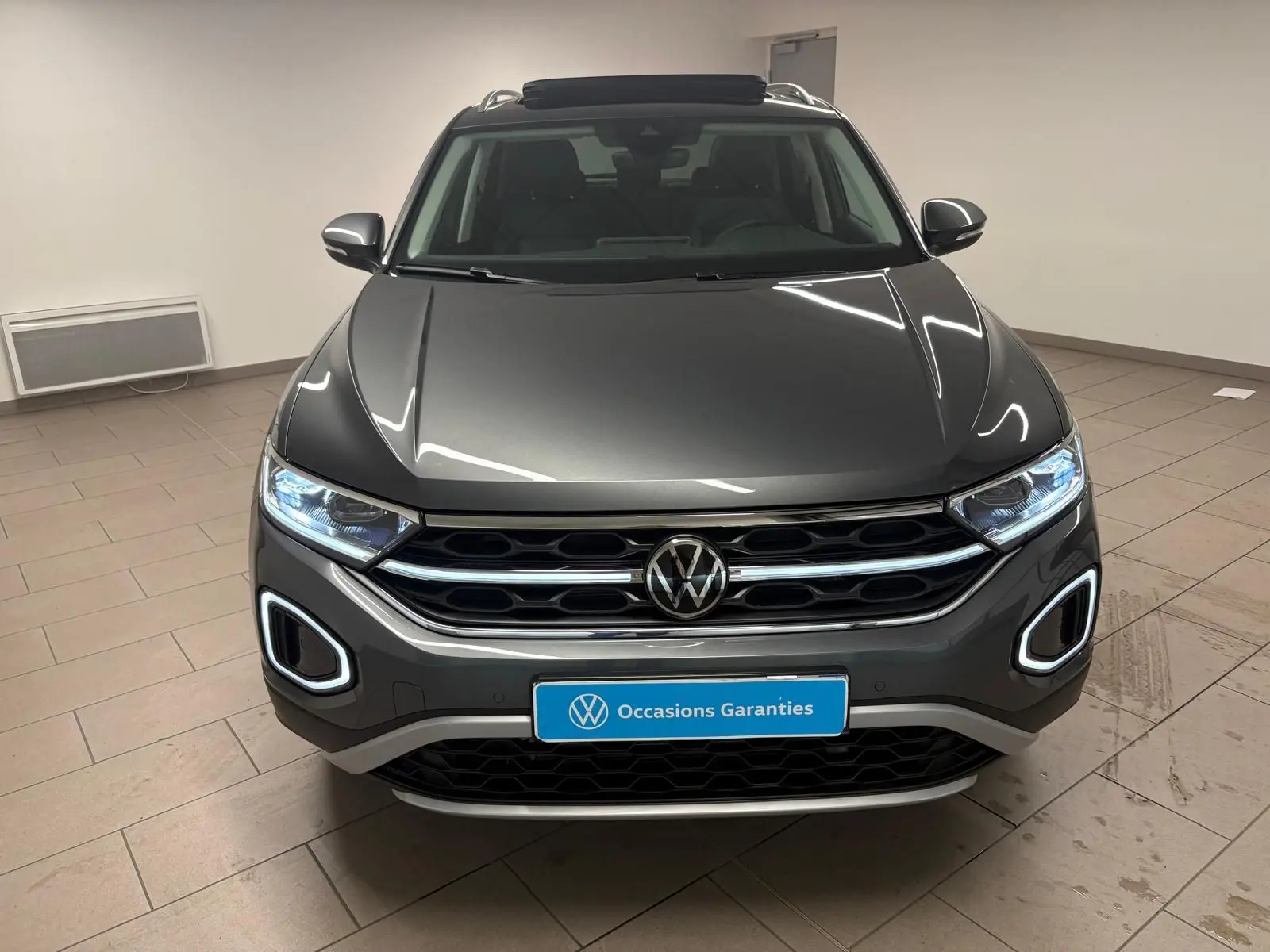 Vue avant d'un Volkswagen T-Roc 2025 gris Indium avec feux allumés et attelage amovible visible en intérieur.