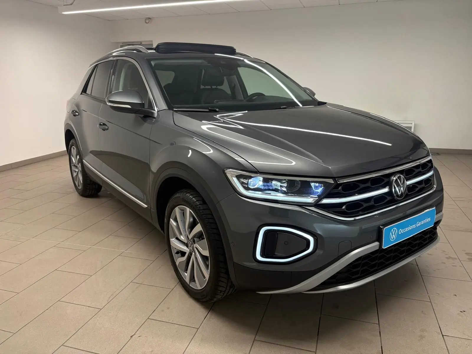 Volkswagen T-Roc 2025 gris Indium en 3/4 avant droit, avec phares LED et attelage amovible visible.