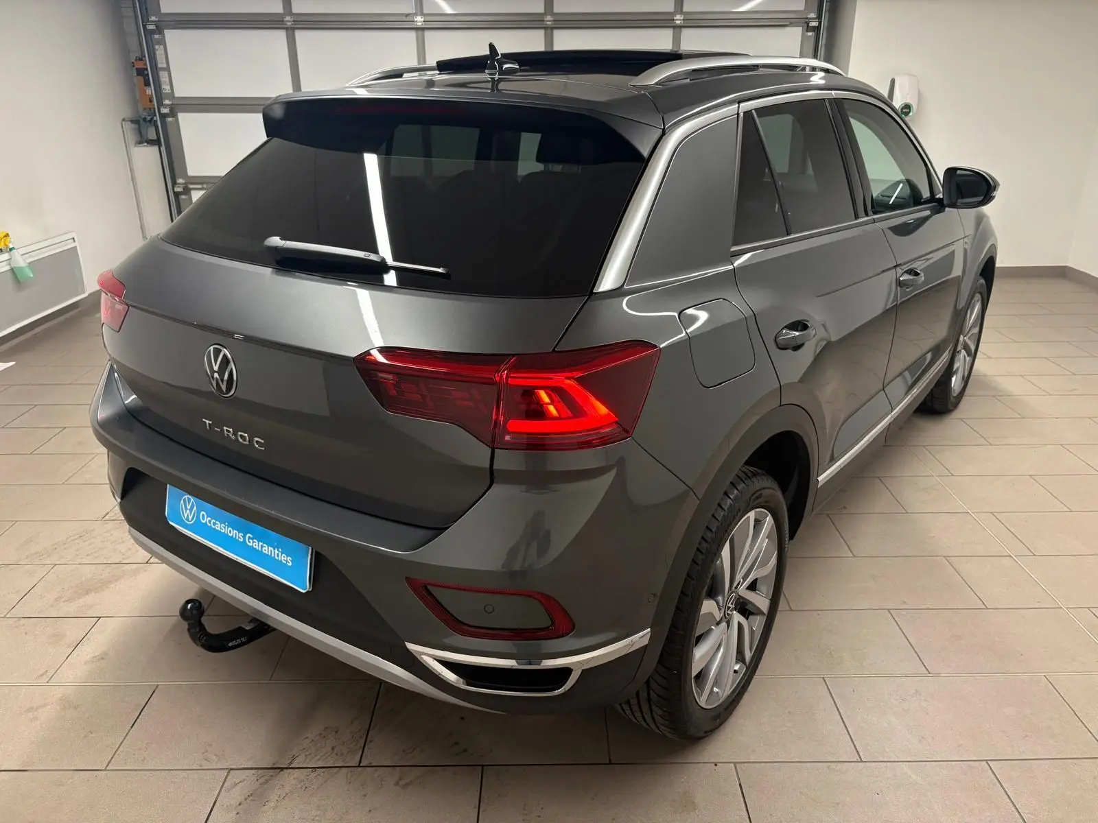 Vue 3/4 arrière droite du Volkswagen T-Roc gris Indium avec attelage amovible et feux arrière allumés en intérieur.