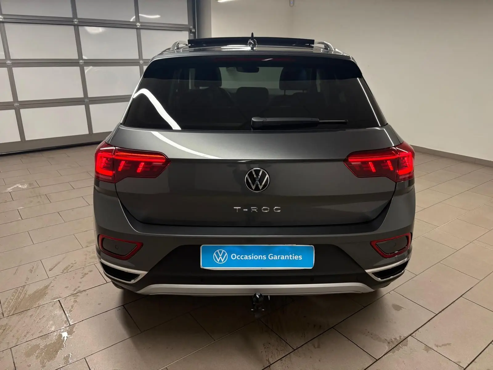 Vue arrière d'un Volkswagen T-Roc gris Indium 2025 avec feux allumés et attelage amovible visible.