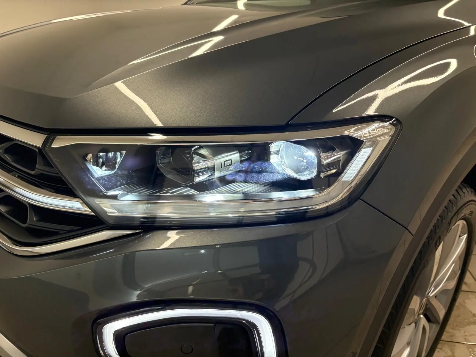 Gros plan sur le phare avant droit du Volkswagen T-Roc 2025 gris Indium avec peinture métallisée et jante visible.