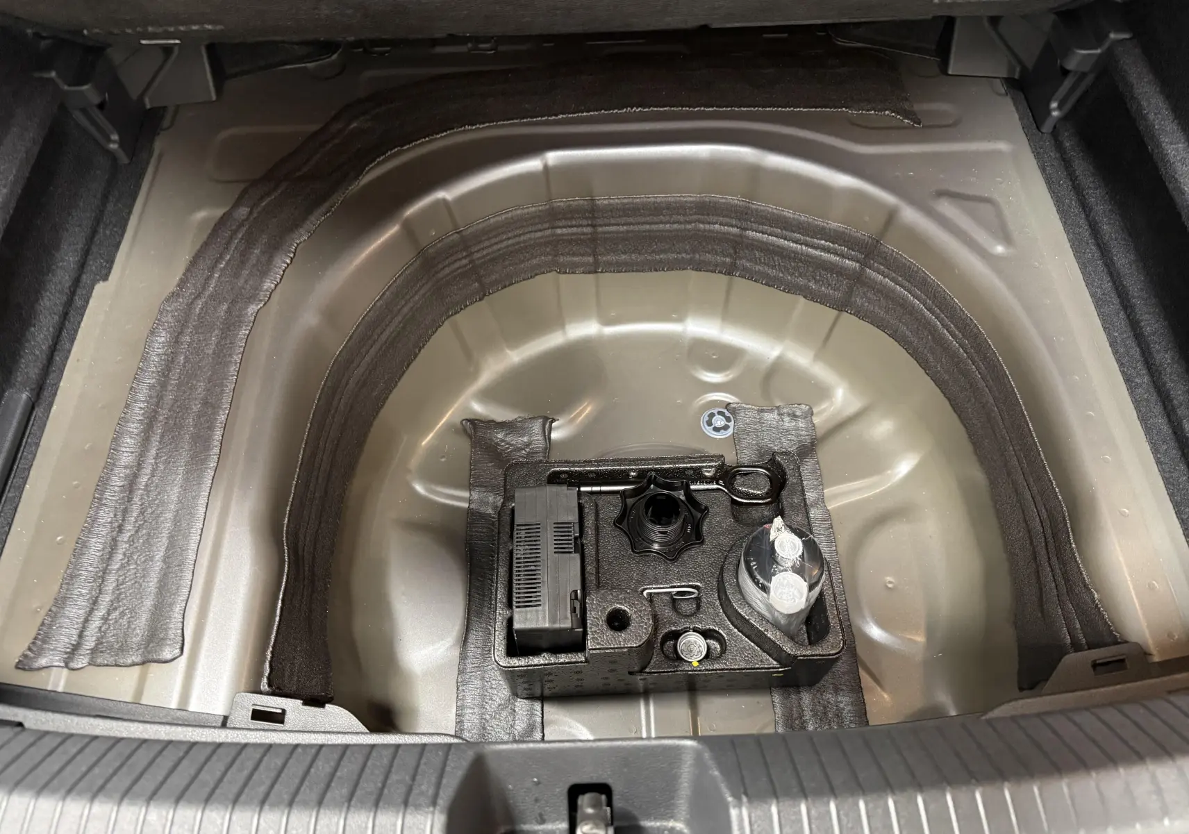 Vue plongeante sur le coffre ouvert du Volkswagen Taigo gris cendre, montrant le rangement pour roue de secours et outils.