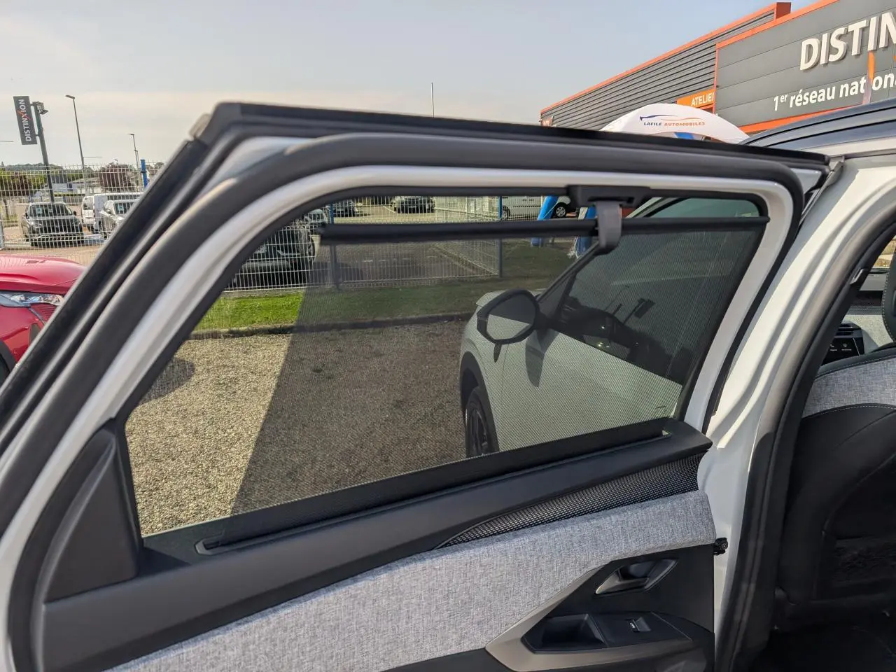 Vue intérieure sur la porte arrière gauche ouverte d’un Peugeot 5008 blanc, mettant en valeur le tissu gris et la protection pare-soleil.