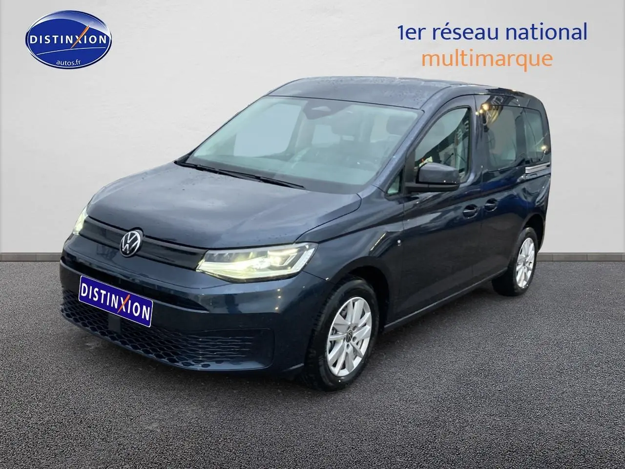 Volkswagen Caddy bleu métallisé vu de 3/4 avant droit, phares allumés sur fond neutre intérieur.