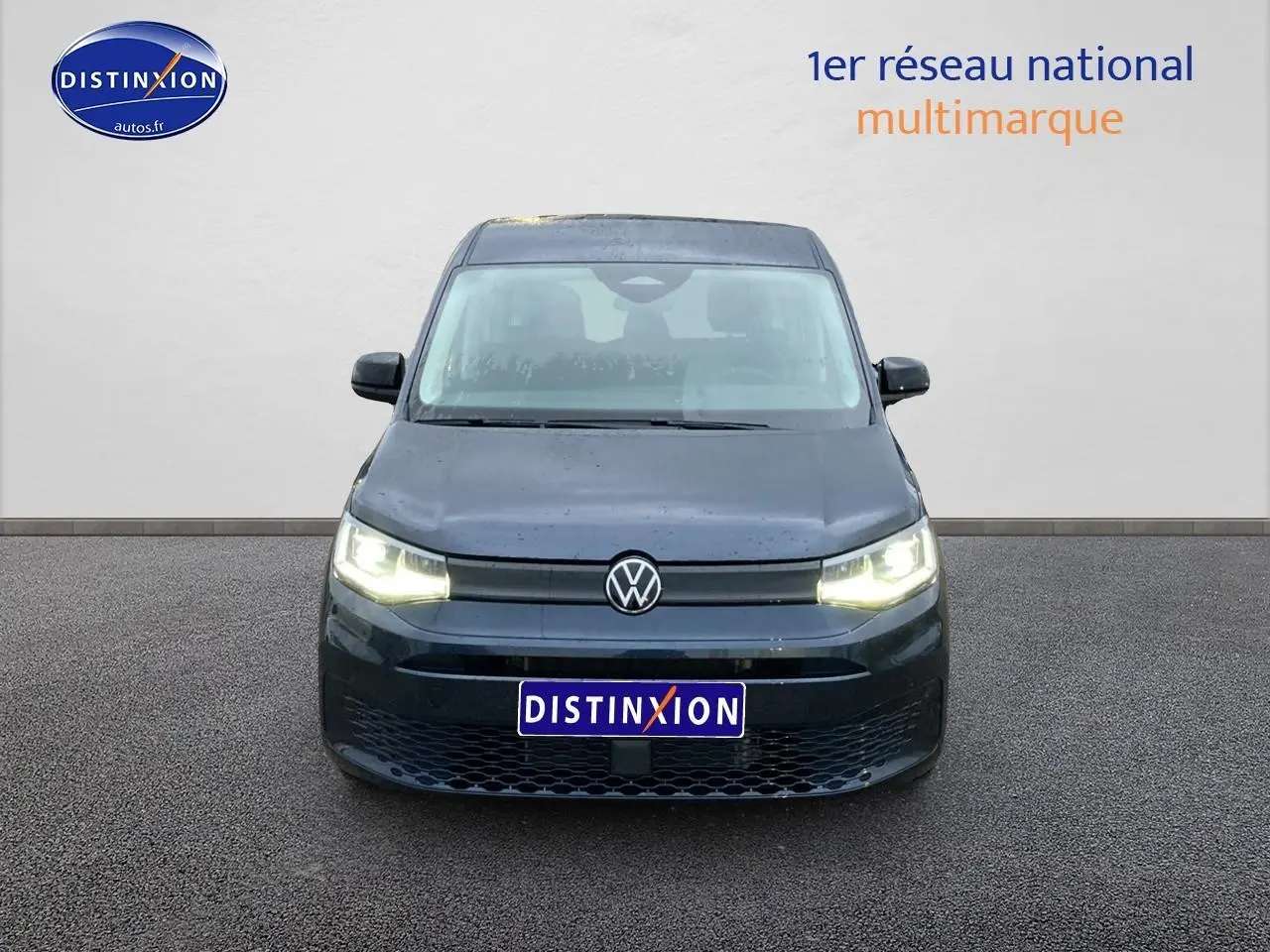 Vue frontale d'un Volkswagen Caddy 2.0 TDI bleu métallisé avec phares allumés sur fond neutre.