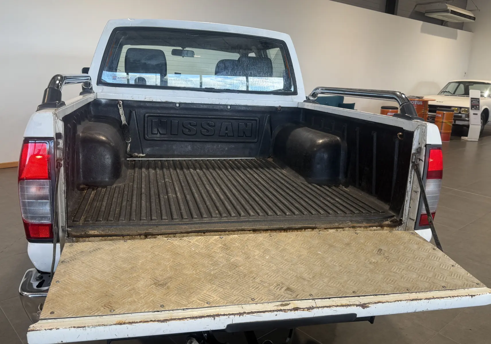 Vue arrière du Nissan Navara NP300 blanc de 2002, plateau ouvert avec benne noire et ridelle abaissée en métal.