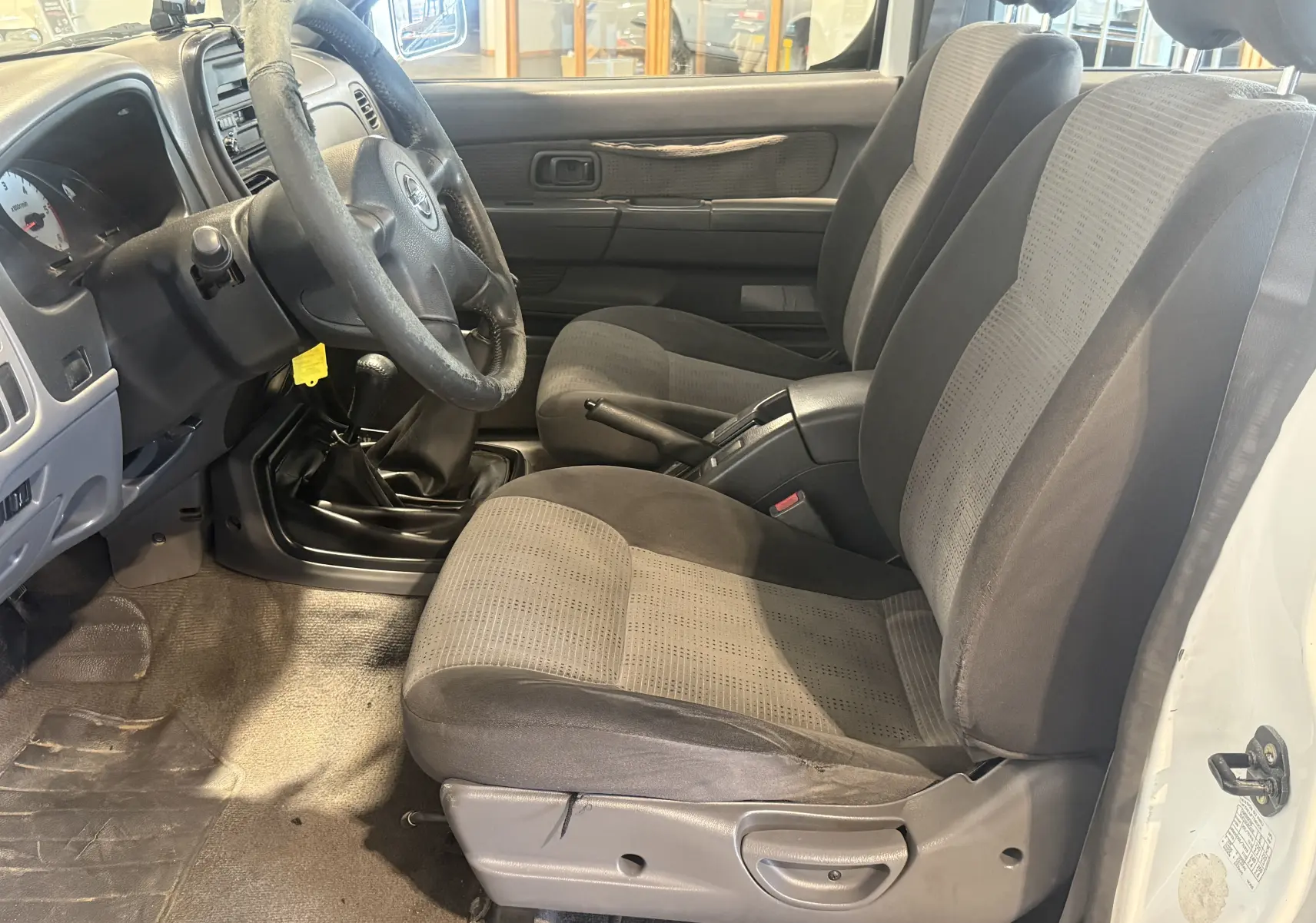 Vue intérieure côté conducteur du Nissan Navara NP300 blanc de 2002, montrant les sièges tissu gris et la boîte manuelle.