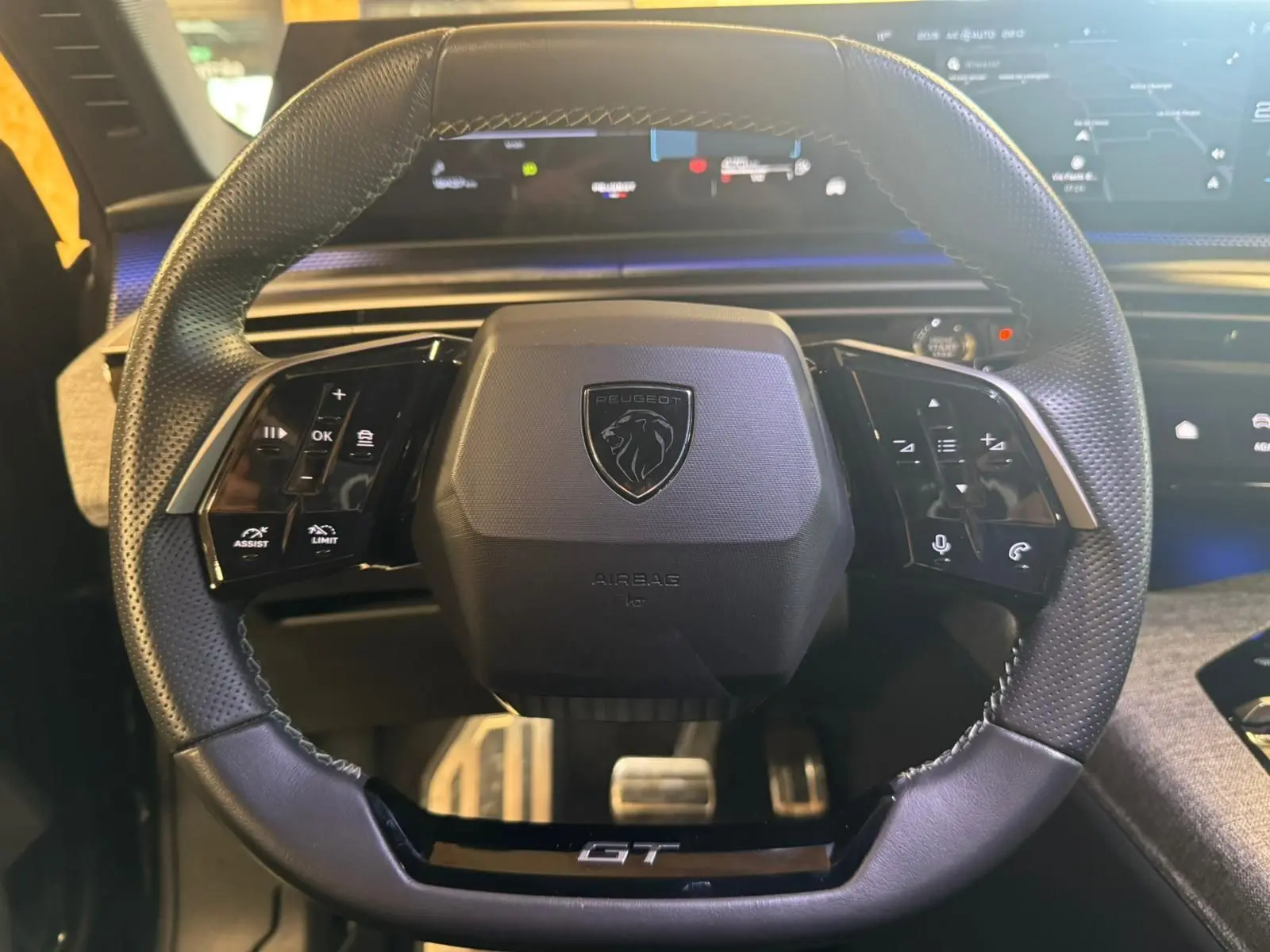 Volant cuir perforé du Peugeot 5008 1.2 HYBRID 136 GT avec logo lion et inscription GT, tableau de bord numérique en arrière-plan.
