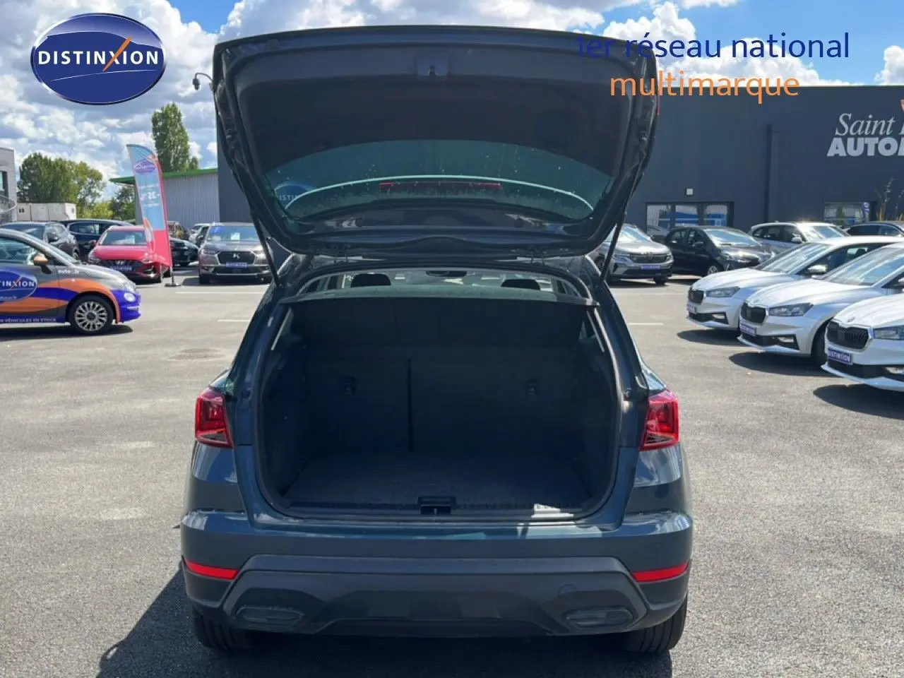 Vue arrière d'un SEAT Arona bleu Fjord avec coffre ouvert, toit blanc visible, sur un parking extérieur ensoleillé.