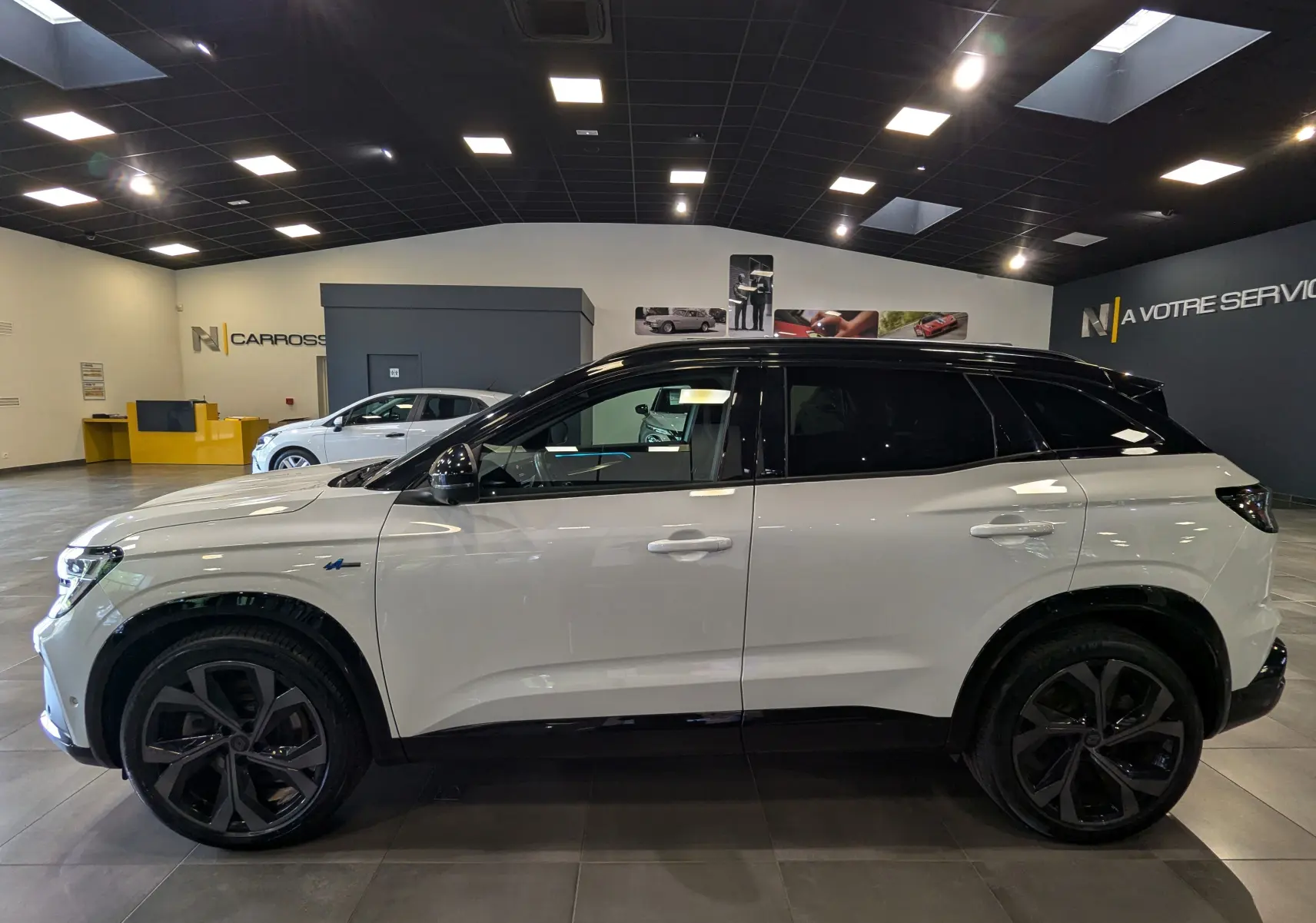 Profil côté gauche du Renault Austral blanc nacré avec toit noir, roues noires et détails Alpine en showroom.