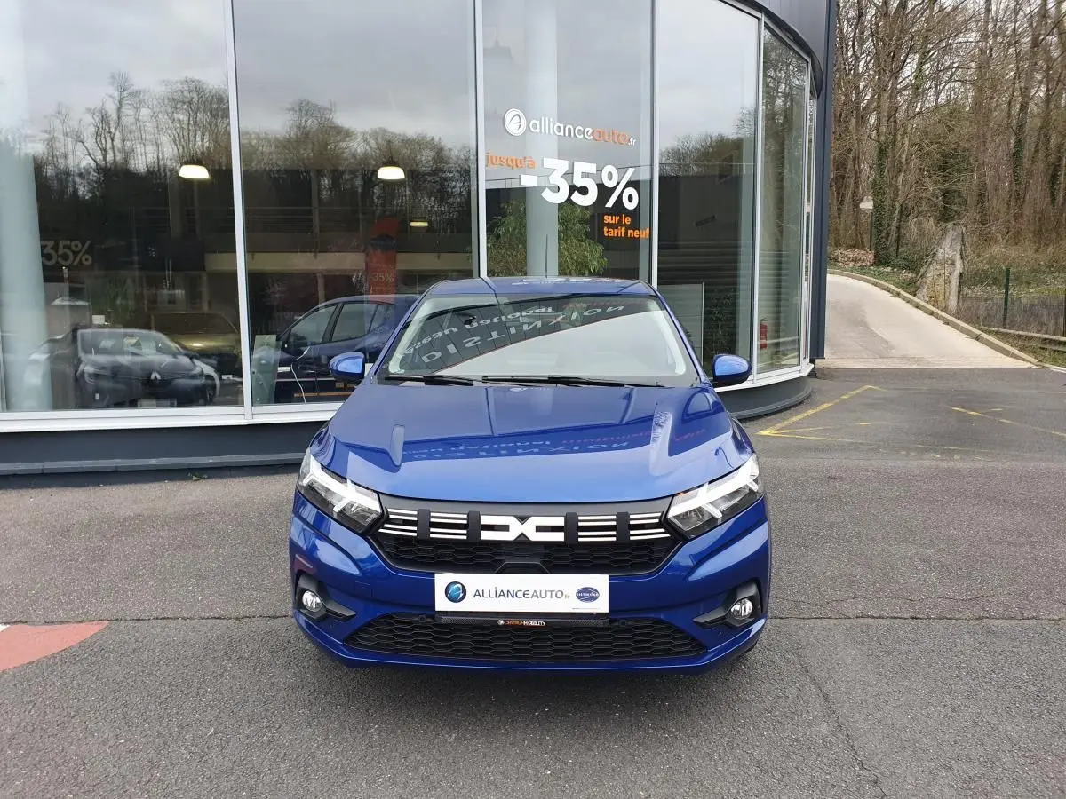 Vue avant d'une Dacia Sandero 2025 bleu Iron avec signature lumineuse LED et feux antibrouillard, stationnée devant un bâtiment.