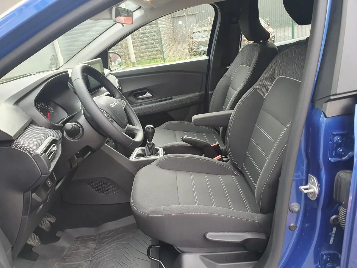 Vue intérieure côté conducteur d'une Dacia Sandero bleu Iron 2025, avec sièges noirs et accoudoir central.