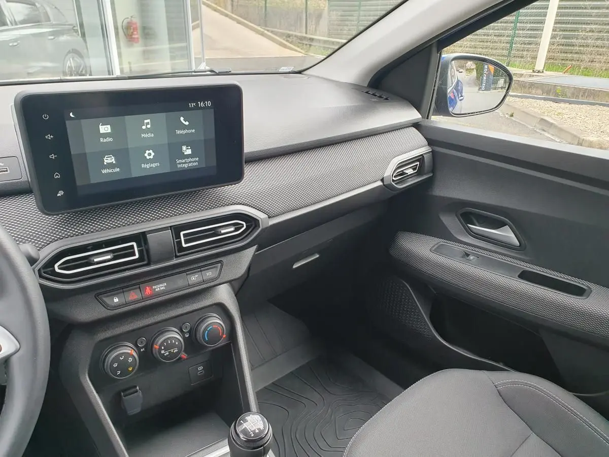 Vue intérieure côté passager du tableau de bord et console centrale de la Dacia Sandero 2025 avec écran tactile et commandes climatisation.