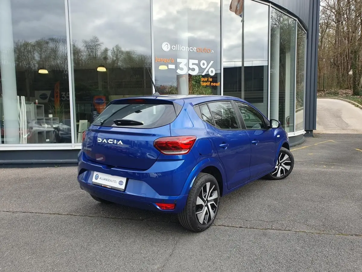 Vue 3/4 arrière droite d'une Dacia Sandero bleu Iron avec enjoliveurs 16'' Atara, stationnée devant un bâtiment vitré.