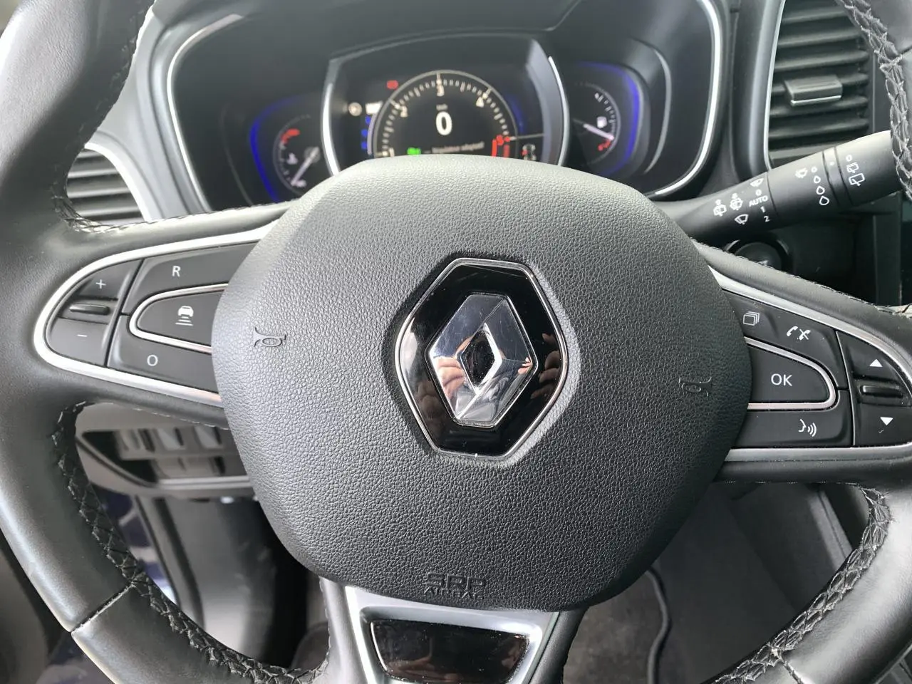 Vue rapprochée du volant en cuir noir avec logo Renault, tableau de bord digital bleu et commandes intégrées visibles.