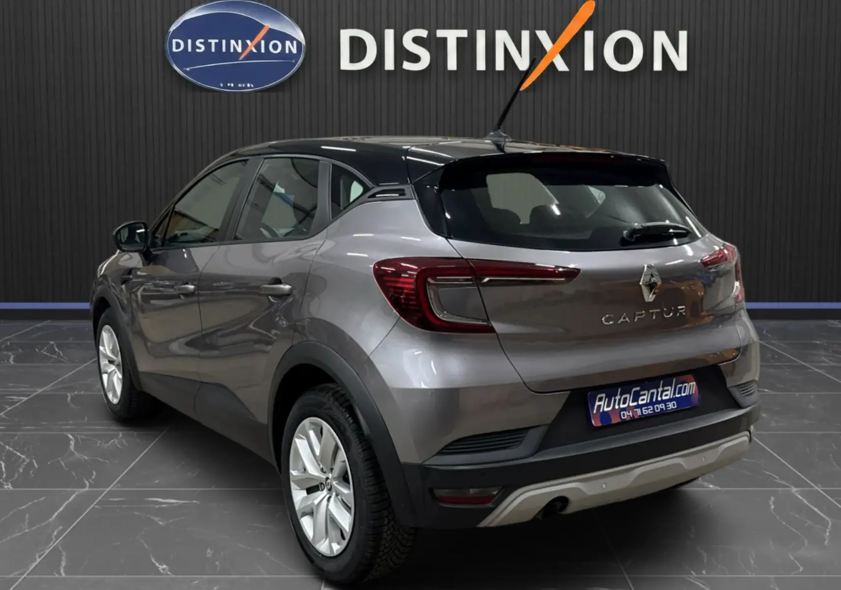 Vue 3/4 arrière droite d'un Renault Captur gris foncé avec toit noir, en intérieur devant un mur noir marqué Distinxion.