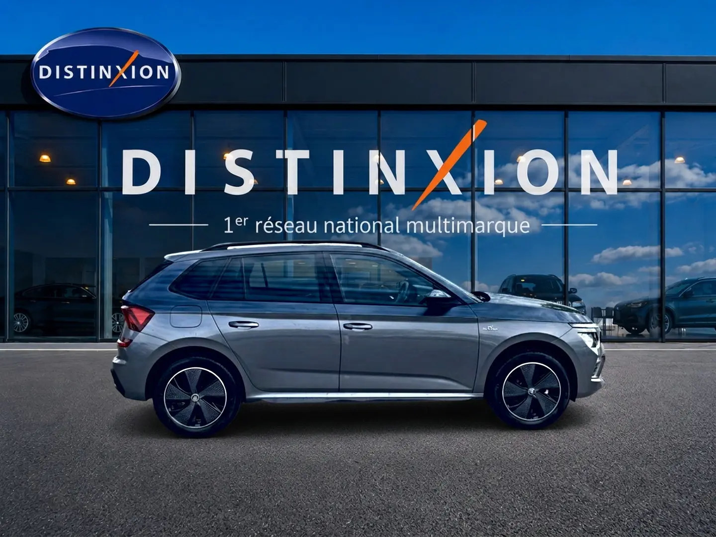 Vue de profil droit du Skoda Kamiq gris graphite avec jantes alu noires et barres de toit visibles devant une vitrine Distinxion.