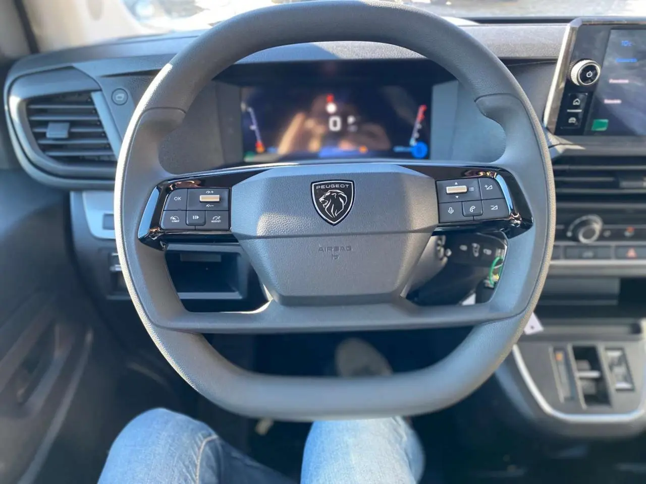 Vue rapprochée du volant Peugeot gris avec commandes intégrées et tableau de bord numérique flou en arrière-plan.