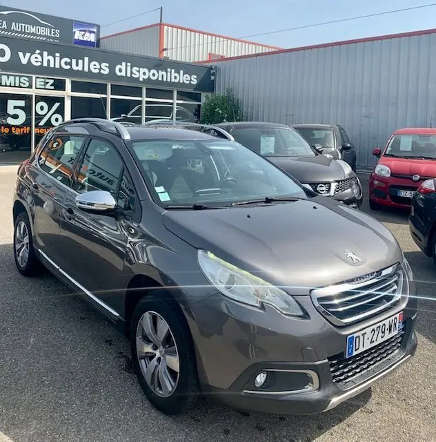Peugeot 2008 gris métallisé vue 3/4 avant droit, avec calandre chromée et barres de toit visibles sur parking.