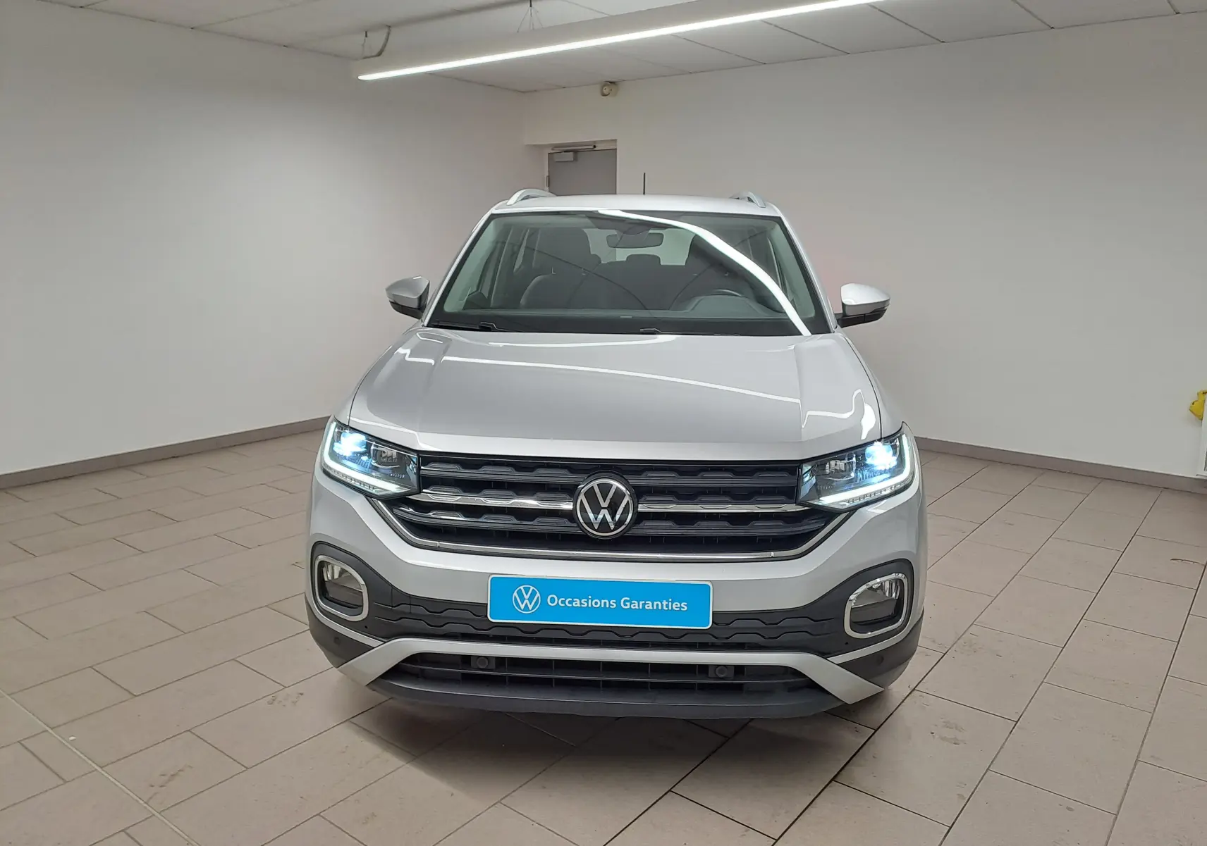 Vue avant d'un Volkswagen T-Cross 1.0 TSI 110 DSG7 Style en peinture métallisée Reflet d'argent avec phares allumés en intérieur.