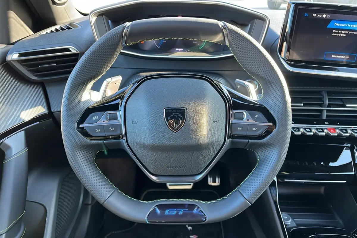 Volant cuir perforé noir avec logo Peugeot et badge GT, tableau de bord numérique et écran tactile central visibles.