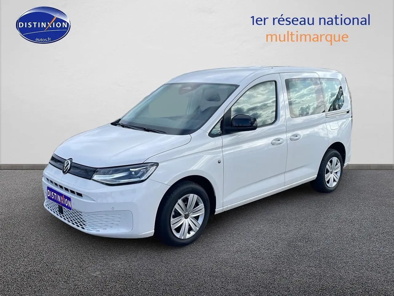 Volkswagen Caddy blanc vue 3/4 avant droit, avec barres de toit et vitres teintées arrière.