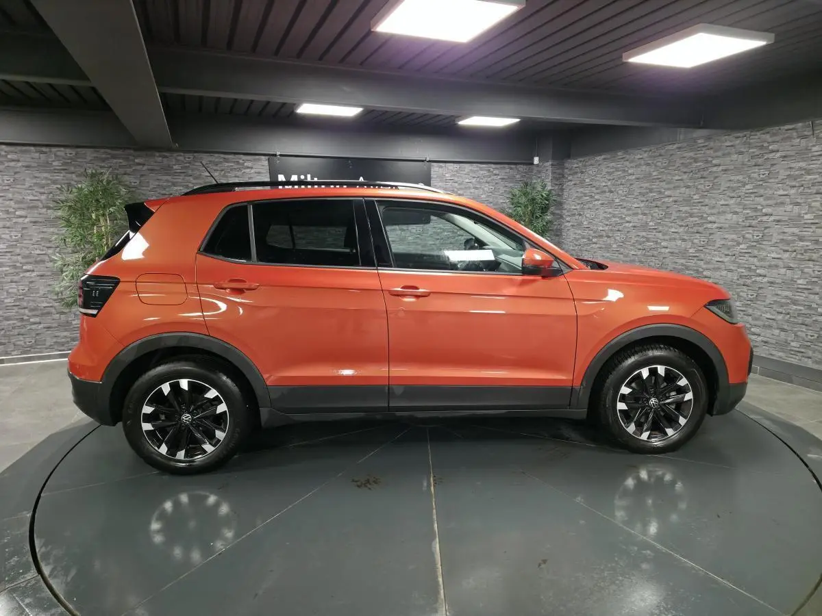 Profil droit d'un Volkswagen T-Cross orange métallisé 2021 avec jantes alliage noires et toit noir en intérieur showroom.