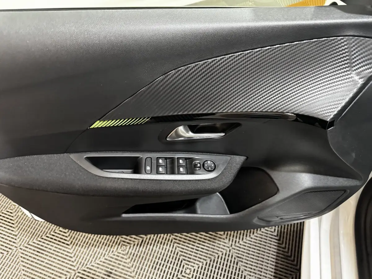 Vue rapprochée de la porte avant gauche noire avec insert effet carbone et commandes électriques sur Peugeot 208 blanc 2024.