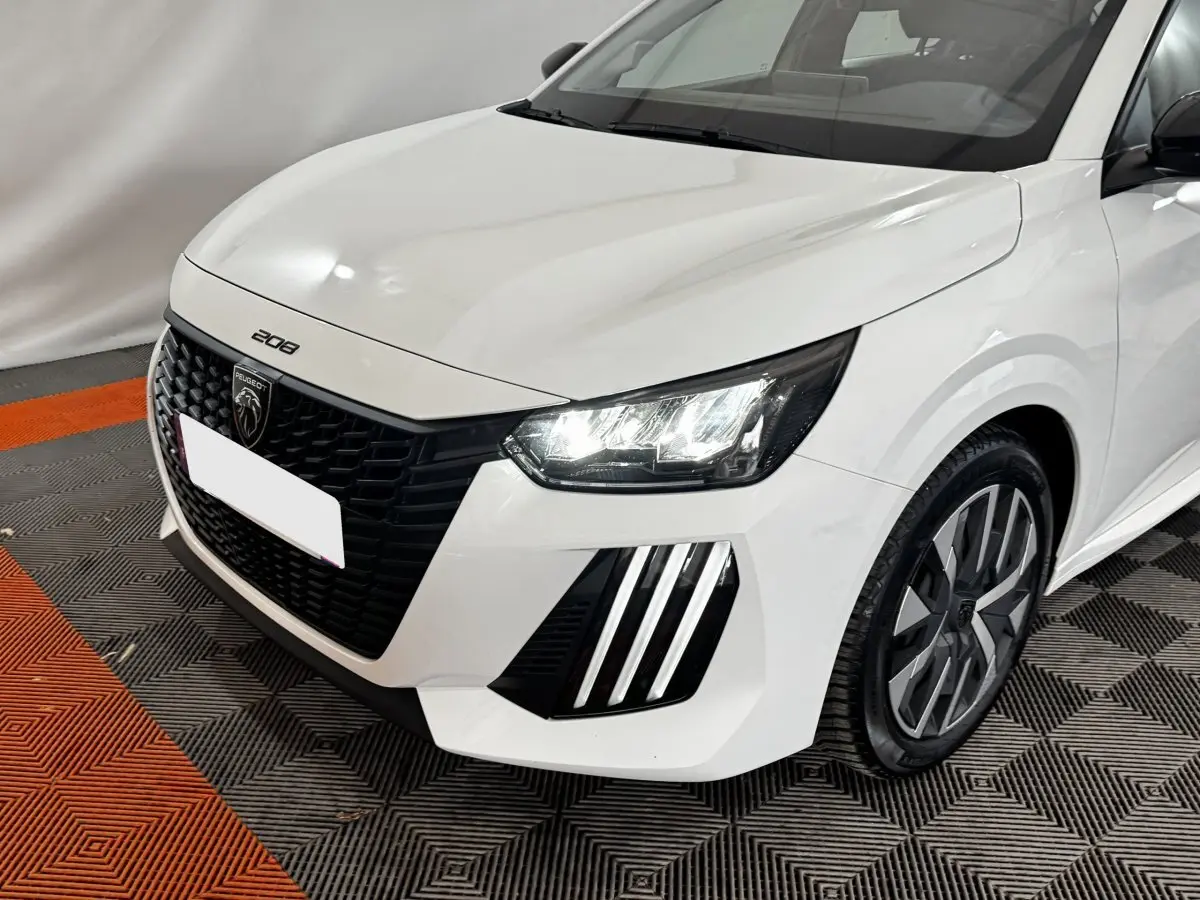 Vue 3/4 avant droite d'une Peugeot 208 blanche avec calandre noire et phares LED allumés.