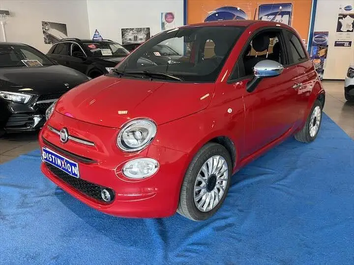 Vue 3/4 avant d'une FIAT 500 rouge brillante avec jantes argentées, exposée en showroom sur tapis bleu.