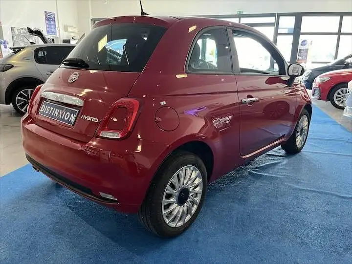 Vue 3/4 arrière droite d'une FIAT 500 rouge hybride exposée en showroom sur un sol bleu.