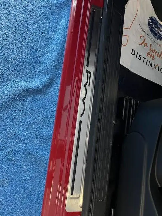 Gros plan sur le seuil de porte côté gauche d'une Fiat 500 rouge 2023 avec logo 500 visible sur fond bleu.