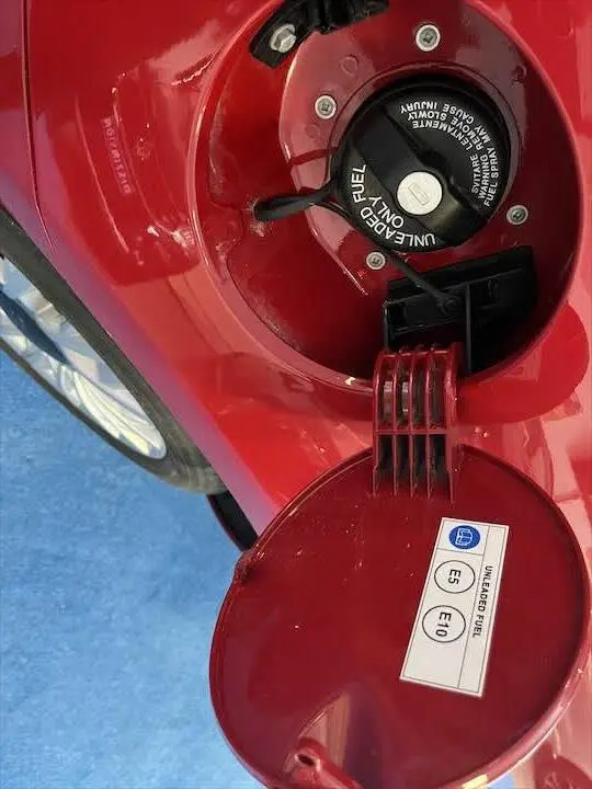 Gros plan sur la trappe à carburant rouge ouverte d'une Fiat 500 2023, avec bouchon essence et étiquette E5 E10 visible.